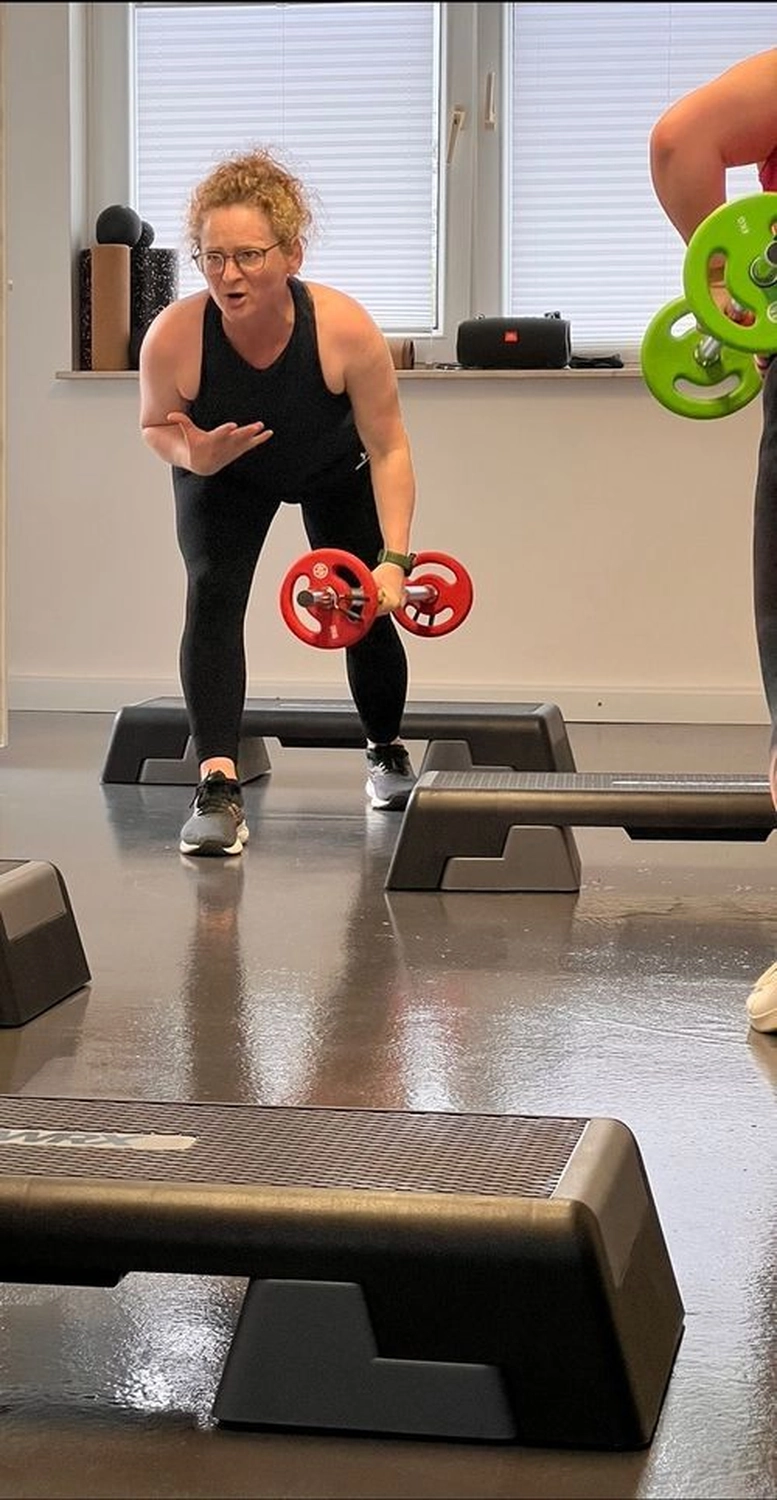 Körpermanufaktur - Physiotherapie. Osteopathie. Naturheilkunde – Dieses Bild zeigt eine Person, die an einem Gruppenfitnesskurs mit Hanteln und Aerobic-Schritten teilnimmt.